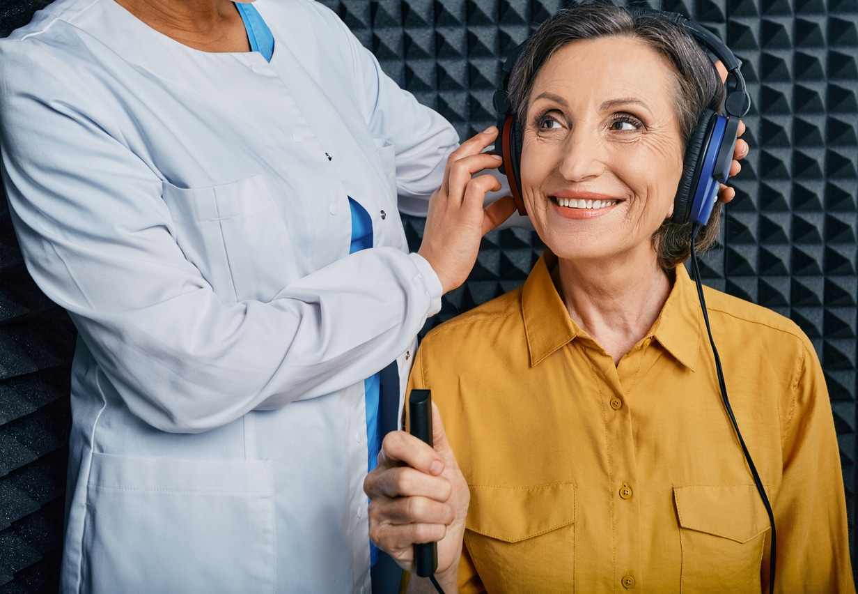A woman sits and has her headphones adjusted by an audiologist while preparing for a hearing test. 